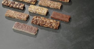 Koyu bir yüzeyde çeşitli enerji çubukları sergilenmiş, granola, çikolata ve meyveli gofretler gibi sağlıklı atıştırmalıklar sergilenmektedir. Karanlık Yüzeyde Çeşitli Enerji Çubukları Sağlıklı Atıştırma Seçenekleri