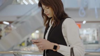 Modern bir alışveriş merkezinde dikilirken akıllı telefonuyla iş kıyafetlerine odaklanmış genç bir kadın. Genç İş Kadını Alışveriş Merkezi 'nde Akıllı Telefon Kullanıyor
