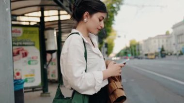 Şehirdeki bir otobüs durağında duran, akıllı telefon kullanan ve toplu taşıma için bekleyen genç bir kadın. Arka planda ağaçlar ve binalar olan gündüz vakti şehir caddesi manzarası. Akıllı Telefon Kullanan Kadın
