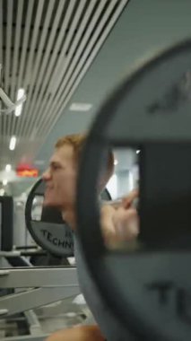 Genç bir adam dayanıklılık eğitimi alıyor, çağdaş bir spor salonunda halter kaldırıyor, fitness ve fiziksel sağlığı vurguluyor. Modern Spor Salonu 'nda Odaklanmış Genç Erkek Ağırlık Kaldıran Ağırlıklar. Dikey video.