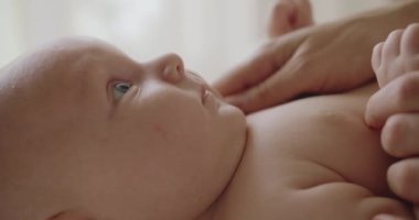 Yumuşak tenli ve sevimli yüzlü çıplak yeni doğmuş bebeğe yaklaş. Anne bebeğini nazikçe okşuyor ve masaj yapıyor. Bebeklik konsepti. Bebek bezli şirin bebek yatakta uzanıp kameraya bakıyor..