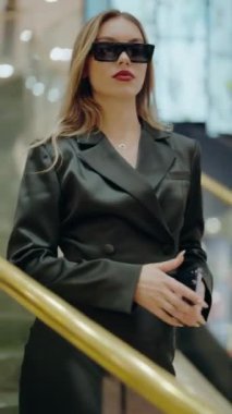 Siyah ceketli ve güneş gözlüklü şık bir kadın, kendine güvenen, modern zarafet ve entelektüellik saçan bir merdivende duruyor. Siyah Ceketli Şık Kadın ve Güneş Gözlüğü İçerde Emin Bir Poz Veriyor