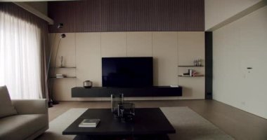 Rahat bir kanepe, ahşap bir medya ünitesi ve zevkli bir minimalist dekor ve büyük parlak pencerelerle çevrili büyük bir düz ekran televizyonu olan modern bir oturma odası..