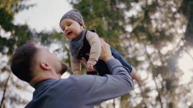 Sevgi dolu bir baba çocuğunu havaya kaldırır, güneşli, ağaçlık bir parkta neşeli bir anı paylaşır. Aile bağlarının ve mutluluğun mükemmel bir sahnesi. Babam neşeli bebeğini dışarı kaldırıyor.