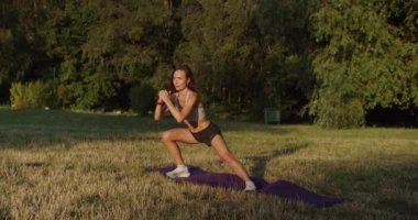 Çekici, zayıf vücutlu, sportif bir kadın. Sağlıklı bir yaşam tarzı ve parkta eğitimi var. Kadın yan hamleler yapıyor, dışarıda egzersiz yapıyor. Genç bir kadın bacak kaldırarak yan hamleler yapıyor..