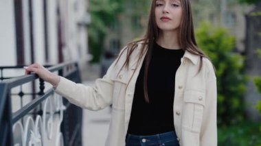 Kendine güveni tam genç bir kadın, günlük kıyafetleriyle şehir sokaklarında poz veriyor. Metal korkulukların yanında rahat bir ifadeyle duruyor, şehir modasını ve rahat bir ruh halini sergiliyor..