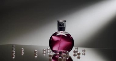 Yansıtıcı bir yüzeyde etrafa saçılmış incilerle çevrili pembe sıvılı şık parfüm şişesi. Ruh hali aydınlatması ayarların zarafetini ve karmaşıklığını vurgular.