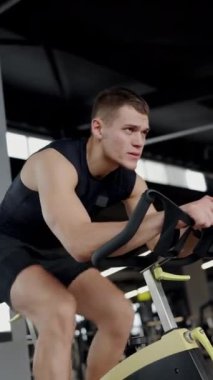 Ciddi bir ifadeyle kararlı genç bir erkek spor salonunda yoğun idman yapıyor. Odaklanma ve zindeliğin özünü yakalar. Odaklanmış Genç Adam Siyah Giyiyor, Sıkı Çalışıyor, Dikey Video.