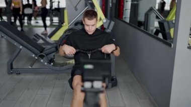 İyi donanımlı bir spor salonunda kürek çeken genç bir erkek azim ve fiziksel zindelik gösteriyor. Genç adam spor salonundaki kürek çekme makinesine odaklandı