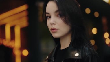 Siyah deri ceketli, siyah saçlı genç bir kadının portresi. Geceleri arka planda bokeh ışıkları olan kameraya bakıyor. Görüntü bir gizem ve içgözlem hissi veriyor..