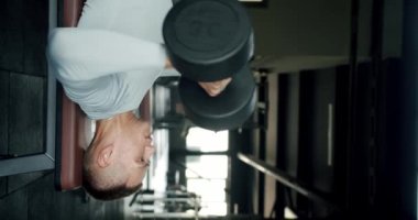 Genç bir erkek sporcu bir bankta uzanmış, halterleri kaldırıyor, modern bir spor salonunda gücünü ve bağlılığını sergiliyor. Spor salonunda Dambıllarla Bench Press 'i sahnelemeye odaklanmış adam. Dikey video.