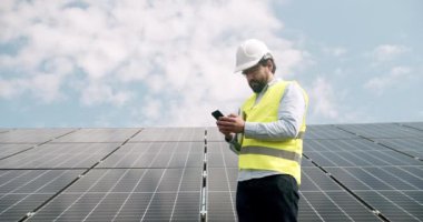 Erkek mühendis, güneş çiftliğinde akıllı telefondan konuşuyor. Düşük açılı üniformalı adam güneş enerjisi istasyonunda güneşli bir günde fotovoltaik panellerin yanında yürüyor ve bulutlu mavi gökyüzüne yapılan çağrıya cevap veriyor..