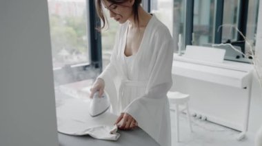 Beyaz bornozlu bir kadın, büyük pencereli ve piyanolu parlak, modern bir odada giysilerini ütülüyor. Sahne sakin ve zariftir. Parlak Modern Daire 'de Giysileri Ütüleyen Kadın