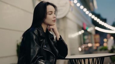 Siyah deri ceketli genç bir kadın gecenin bir yarısı açık bir kafede oturuyor, düşüncelere dalmış. Sokak ışıklarla süslenmiş, sakin ve yansıtıcı bir atmosfer yaratıyor..
