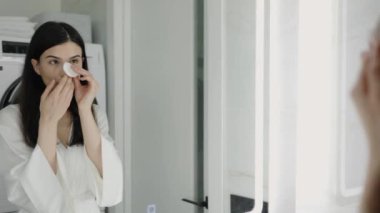 Beyaz bornozlu genç bir kadın banyoda duruyor, yüzünü temizlemek için pamuk ped kullanıyor. Parlak ayna sakin, yansıtıcı bir atmosfer yaratır..