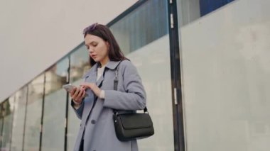 Modern bir ofis kompleksinin dışında akıllı telefonunu kullanan şık trençkotlu profesyonel genç bir kadın, bağlantı ve şehir yaşam tarzını tasvir ediyor. Genç İş Kadını Akıllı Telefon Kullanıyor