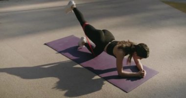Sportif kadın dışarıda egzersiz minderinde çalışıyor. Spor kıyafetli terli kadın uyluk kasları üzerinde egzersiz yapıyor, bacağını kaldırıyor ve indiriyor, kalçasını geriyor, spor lastiği kullanıyor..