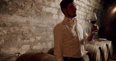 Mutlu erkek şarap üreticisi bir lezzet tadıyor ve şarap mahzenindeki şeffaf bardaklara dökülen taze kırmızı şarap kalitesini kontrol ediyor. Güzel giyimli Sommelier bir bardak kırmızı şarabı kontrol edip içiyor.