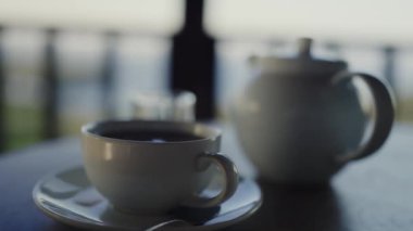Bir kahve fincanı, çaydanlık ve şeker kabının olduğu rahat bir sabah sahnesi, güne barışçıl bir başlangıç olduğunu gösteriyor. Bardaklı Sabah Kahve, Çaydanlık ve Masada Şeker