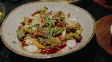 Mantarlar, biberler ve yeşilliklerle dolu, siyah bir masada seramik bir kasede servis edilen gurme sebze kızartmasının yakın çekimi. Görsel olarak çekici ve iştah açıcı bir vegan yemeği..