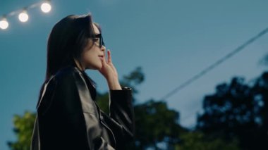 Deri ceket ve güneş gözlüğü takan kendine güveni tam bir kadın, dışarıda tel ışıkların altında duruyor. Modern, şık bir görüntü, özgüven ve akşam havası sergiliyor. Güneş gözlüğü takan şık bir kadın