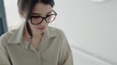 Gözlüklü genç bir kadın minimalist ofis ortamında dizüstü bilgisayarıyla çalışıyor. Konsantrasyon, verimlilik ve modern çalışma alanı. Kapalı alanda çalışan odaklanmış gözlüklü genç bir kadın.