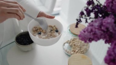 Bir el, bir kase tahıl gevreğini süt ve dutla tutarken, besleyici bir kahvaltıyı sembolize ediyor. Bir kavanoz granola ve bir kase yaban mersini de görülebilir..