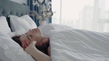 Bir kadın, bembeyaz çarşaflarla kaplı, yumuşak, doğal sabah ışığıyla aydınlanmış bir şekilde yatakta huzur içinde yatar. Sakinleştirici atmosfer, sakin ve rahat bir yatak odası ortamında huzur ve rahatlık olduğunu gösteriyor..