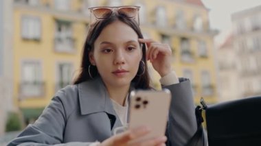 Genç, rahat bir kadın açık bir kafedeki masada oturuyor ve akıllı telefonunu kullanırken gülümsüyor. Renkli binalar canlı bir zemin oluşturur. Kentte Akıllı Telefon Kullanırken Kahve İçen Genç Kadın