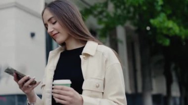 Genç bir kadın dışarıda eğleniyor, akıllı telefonunu kullanıyor ve elinde bir fincan kahve tutuyor. Mutlu ve rahat görünüyor. Elinde kahve fincanı tutarken dışarıda akıllı telefon kullanan genç bir kadın.
