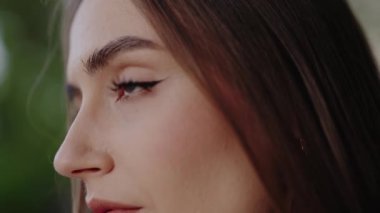 Güzel makyajlı, gözlerini ve doğal güzelliğini ön plana çıkaran bir kadının yan görüntüsü. Görüntü bulanık yeşil arka planda sakin ve sakin bir ifade yakalıyor. Kadın Yüzü
