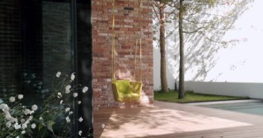 Modern bir havuz kenarında asılı, beyaz çiçekler ve tuğla duvarla tamamlanmış canlı sarı bir rattan sandalyeyi gösteren sakin bir açık hava sahnesi. Modern Ev Bahçesi