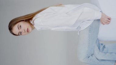 Sıradan beyaz gömlekli, kot pantolonlu, minimalist bir stüdyo ortamında oturan ve poz veren zarif genç bir kadın. Bakışları düşünceli, sakin bir ifadeyle. Beyaz Gömlekli Genç Kadın. Dikey video.