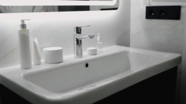 Modern bir banyo lavabosu ve mermer tezgahta sergilenen parlak cilt ürünleri. Temiz ve minimalist tasarım bir lüks ve basitlik hissi yayar. Modern Banyo Lavabosu