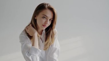 Beyaz tişörtlü genç kadın, gündelik pozlar veriyor. Beyaz gömlekli, uzun saçlı genç bir kadının portresi. Hafif aydınlık bir ortamda sakin ve huzurlu bir ifade yayıyor..
