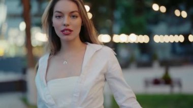 Gece boyunca açık havada duran beyaz elbiseli zarif genç bir kadın. Arka planda hafifçe parlayan parlak, dekoratif ışıklar. Kendine güven ve güzellik saçıyor. Portre Genç Güzel Kadın