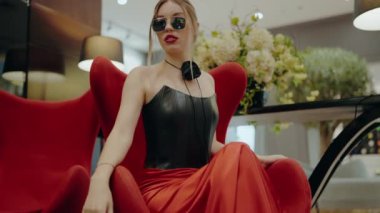 Moda sahibi bir kadın, modern, zarif bir iç mekanda, güneş gözlüğü ve siyah bir elbise takan kırmızı bir sandalyeye güven içinde oturur. Kırmızı Sandalyedeki Şık Kadın Güneş Gözlüğü ve Siyah Elbise Giyiyor 