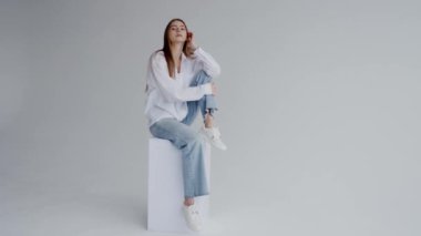 Rahat beyaz gömlek ve kot pantolon giymiş, sade bir arka plana yaslanmış, sakin ve cana yakın bir tavır sergileyen genç bir kadının portresi. Günlük Kıyafet Pozisyonunda Genç Kadın