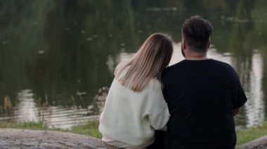 Sakin bir gölün kenarında oturan bir çiftin romantik görüntüsü. Aşk, ilişkiler ve dinginlik teması için mükemmel. Romantik Çift Huzurlu Göl Manzarasının Tadını Çıkartıyor