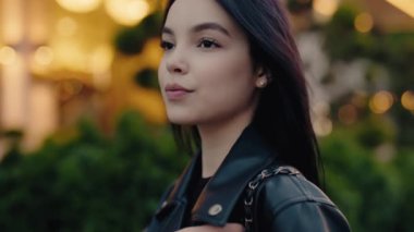 Kendine güveni tam genç bir kadın deri ceket giyiyor ve alacakaranlıkta dışarıda poz veriyor. Arka planda bulanık ışıklar var. Deri ceketli kendine güvenen genç kadın Dusk 'ta