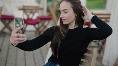Genç bir kadın açık bir kafede selfie çekiyor, rahat ve rahat görünüyor. Oturmuş ve siyah bir bluz ve kot pantolon giymiş, sakin atmosferin tadını çıkarıyor. Genç Kadın Selfie Çekiyor