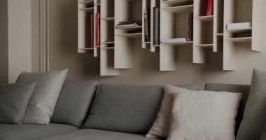 Modern bir oturma odası. Duvara monte edilmiş çeşitli kitaplarla donatılmış şık bir kitaplık ve yastıklarla süslenmiş rahat gri bir kanepe. Rahat ve davetkar bir ortam yaratıyor..