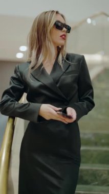 Siyah elbiseli ve güneş gözlüklü şık bir kadın şehir ortamında yürürken kendine güvenir. Modern şehir sahnesinde moda, zarafet ve cesaret tasvir edilir. Moda Kadını