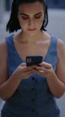 Genç bir kadın dışarıda telefonunu kullanıyor. Kayarken ve yazarken sakin ve odaklanmış görünüyor. Şık bir saç kesimi var ve şık kıyafetler giyiyor. Şehir Merkezinde Şık Kadın Pozisyonu,
