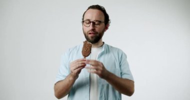 Gözlüklü ve sakallı bir adam beyaz arka planda çikolata kaplı bir dondurmanın tadını çıkarıyor. Bir ısırık alır, gözlerini kapatır ve mükemmel bir yaz ziyafeti sahnesi yaratarak saf zevk gösterir..