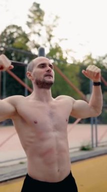 Kaslı bir adam bisepslerini açık hava spor sahasında gösterir. Üstsüz ve siyah şort giyiyor. Derin nefes alıyor, kaslı fiziğini ve sıkı çalışmasının sonuçlarını gösteriyor..