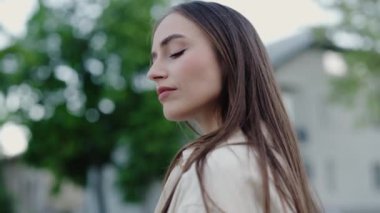 Uzun kahverengi saçlı, kendine güvenen genç bir kadın, bir bahar günü dışarıda duruyor, kararlı görünüyor. Arka planda Yeşil Yapraklı Serene Genç Kadın