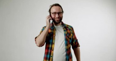 Gözlüklü ve sakallı neşeli bir adam konuşurken jestler kullanarak animasyonlu bir telefon görüşmesi yapıyor. Renkli bir ekose gömlek giyiyor, basit beyaz bir arkaplanda duruyor..