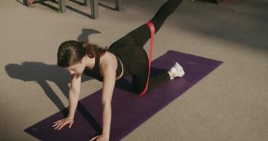 Yoga tahtası pozu veren bir kadın. Spor kıyafetli terli kadın uyluk kasları üzerinde egzersiz yapıyor, bacağını kaldırıyor ve indiriyor, kalçasını geriyor, spor lastiği kullanıyor..