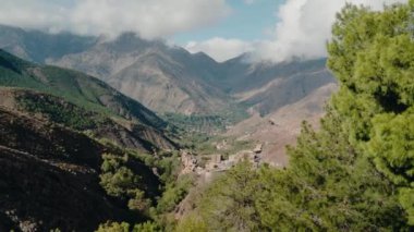 Atlas Dağları arasında bir vadiye kurulmuş Fas köyünün manzarası. Yeşiller yemyeşil ve geleneksel evler sakin bir atmosfer yaratıyor. Bulutlar tepelerin üzerinde sürükleniyor,